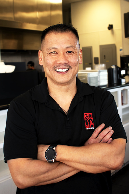 Image contains: Asian Male with a big smile and wearing KoJa Kitchen shirt with interior of a restaurant in background
